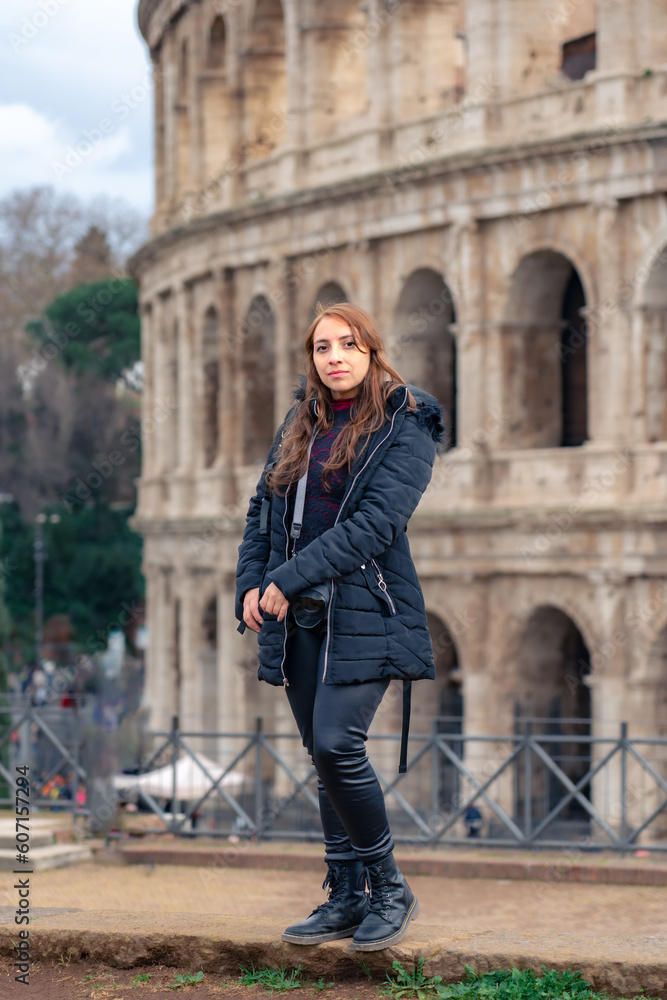 Pensive young Latin woman tourist in warm clothes and professional camera standing against Colosseum and looking away while admiring historic architecture during trip through Rome, Italy
