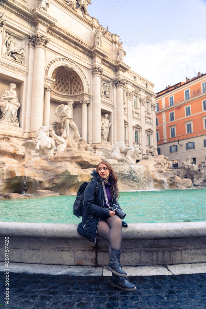 Content young Latin woman tourist in warm clothes sitting on border of Trevi Fountain with professional camera and looking away while enjoying amazing architecture in Rome, Italy during vacation