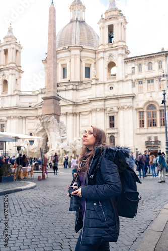 Trendy young Latin woman tourist in sunglasses and warm clothes standing with professional photo camera looking away in Piazza Navona in Rome, Italy