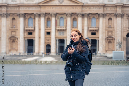 Happy young Latin woman in warm clothes using her smartphone and professional photo camera while standing near historic church Saint Peter Basilica in Vatican city, Rome, Italy 