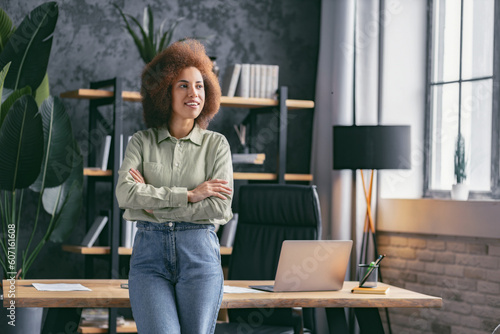 Portrait of young happy confident African American business woman executive. Successful female entrepreneur standing near a desk in office looking away and smiling