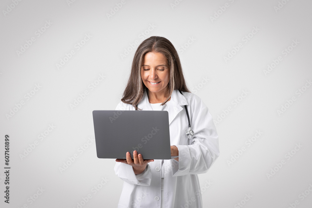 Happy confident european senior woman doctor in white coat with stethoscope using laptop on light gray background Happy confident european senior woman doctor in white coat with stethoscope using laptop on light gray background