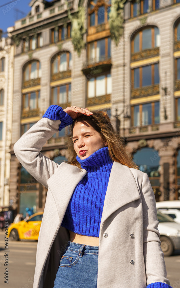 portrait of a brunette girl in coat and bright blue sweater on background of a busy city street. bright sun, hard shadows. concept of a real person, not a model. walking down the street, spring mood