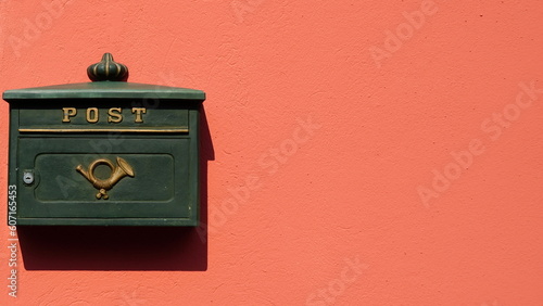 Green metal mailbox with golden post symbol on bright orange plastered wall in the sunshine. Space for text. 