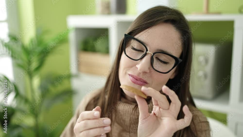 Beautiful hispanic woman eating cookie sitting on table at dinning room Beautiful hispanic woman eating cookie sitting on table at dinning room