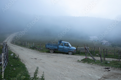 A village in foggy weather. A man walking on village path. There is a blue truck.