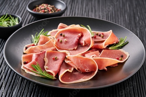 parma ham prosciutto with rosemary and spices in a plate on a black wooden table.