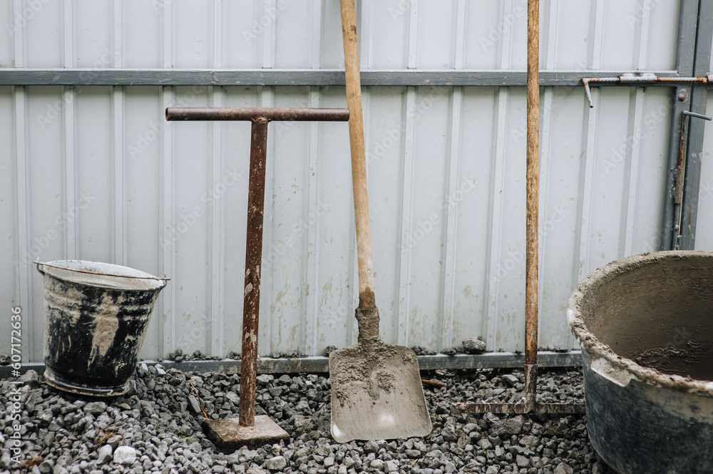 Construction tools: a bucket, a shovel, a rake, a concrete mixer in concrete, cement stand on stones against the background of a house wall. Construction tools: a bucket, a shovel, a rake, a concrete mixer in concrete, cement stand on stones against the background of a house wall.