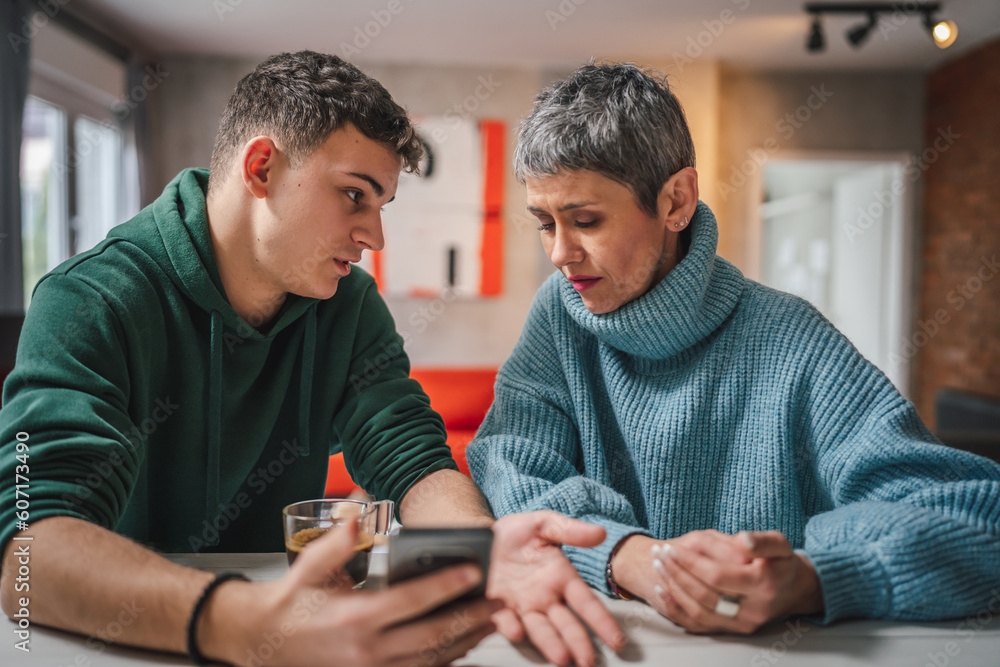 teenage boy and mature caucasian woman sit together at the kitchen at home talk mother and son or relatives support solving problem share experience and opinion boy hold mobile phone smartphone teenage boy and mature caucasian woman sit together at the kitchen at home talk mother and son or relatives support solving problem share experience and opinion boy hold mobile phone smartphone