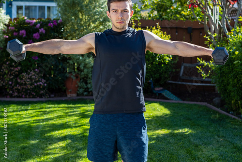 Young adult male doing lateral raises to target his shoulders during an afternoon at-home workout on a yoga mat during the coronavirus pandemic.