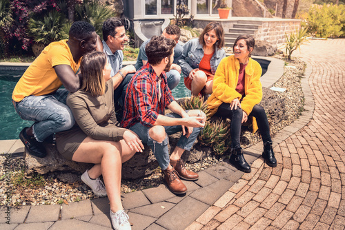 A diverse group of young people, aged 20-40, is lounging by a pool in a courtyard. They're chatting, dressed in spring attire, and enjoying each other's company. photo