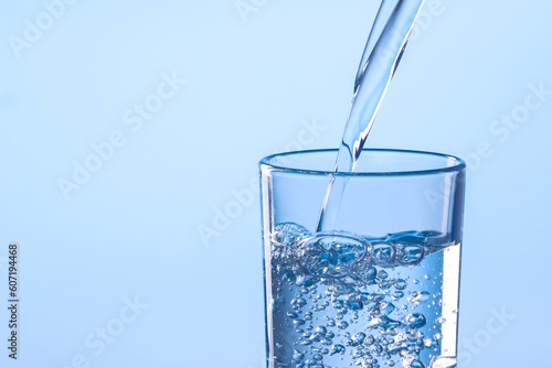 Pouring water into glass against light blue background, closeup. Space for text