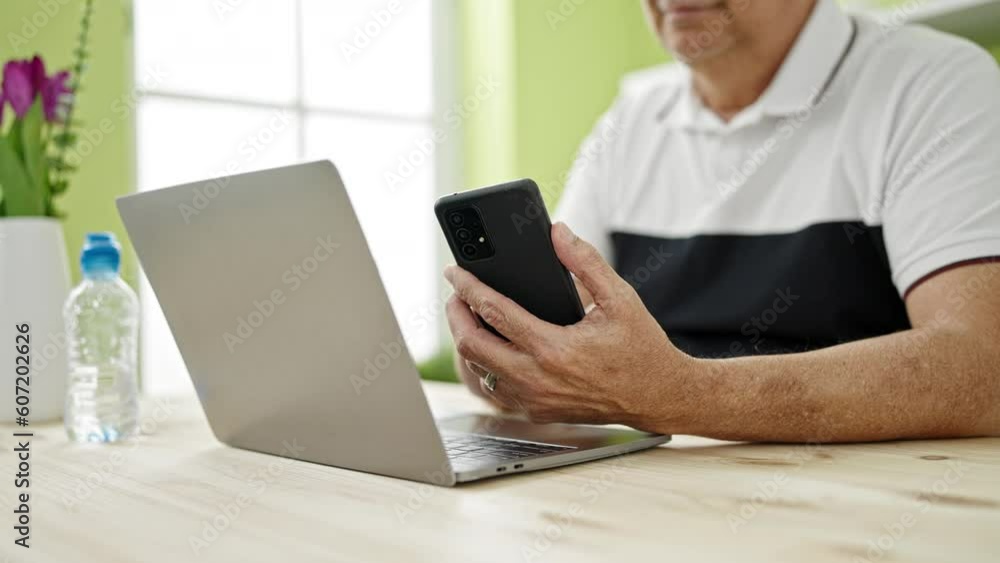 Middle age man with grey hair using smartphone and laptop at dinning room