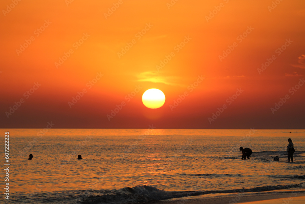Por do sol em tons de laranja na beira do mar em ferias de verao com pessoas na agua.