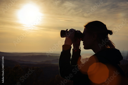 A young adult female hiker and traveler looks through binoculars in the mountains in the magical evening light of a sunset © Kate Stock