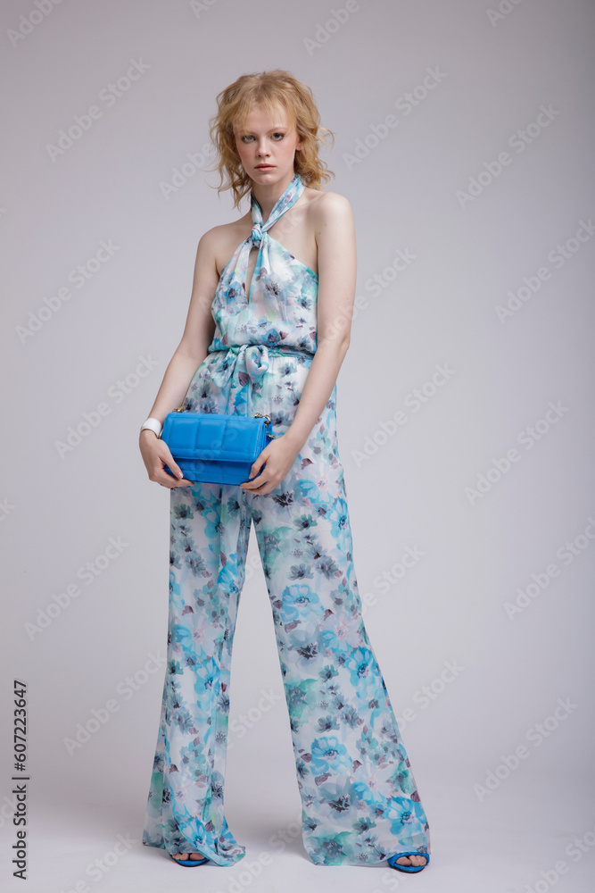 High fashion photo of beautiful elegant young woman in a pretty rich blue jumpsuit with floral pattern, handbag, clutch posing over white, soft gray background. Studio Shot, portrait. Blonde High fashion photo of beautiful elegant young woman in a pretty rich blue jumpsuit with floral pattern, handbag, clutch posing over white, soft gray background. Studio Shot, portrait. Blonde