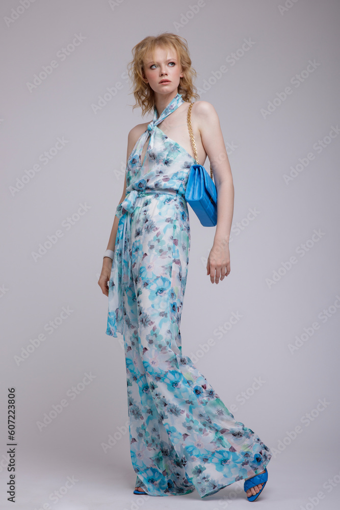 High fashion photo of beautiful elegant young woman in a pretty rich blue jumpsuit with floral pattern, handbag, clutch posing over white, soft gray background. Studio Shot, portrait. Blonde High fashion photo of beautiful elegant young woman in a pretty rich blue jumpsuit with floral pattern, handbag, clutch posing over white, soft gray background. Studio Shot, portrait. Blonde