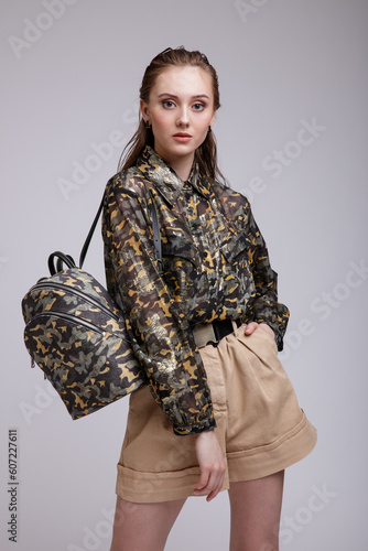 High fashion photo of beautiful elegant young woman in a pretty military camouflage shirt, sneakers, backpack, beige shorts posing over white, soft gray background. Studio Shot, portrait.