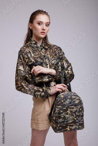 High fashion photo of beautiful elegant young woman in a pretty military camouflage shirt, sneakers, backpack, beige shorts posing over white, soft gray background. Studio Shot, portrait.