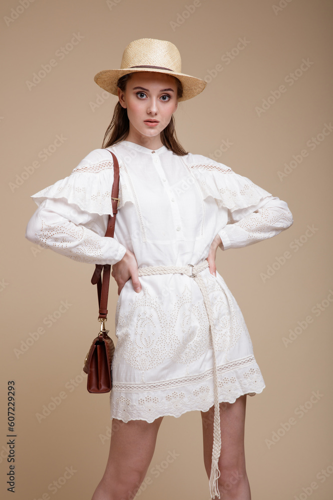High fashion photo of beautiful elegant young woman in a pretty white dress with the sleeves, hat, handbag posing over beige brown background. Studio Shot, portrait. High fashion photo of beautiful elegant young woman in a pretty white dress with the sleeves, hat, handbag posing over beige brown background. Studio Shot, portrait.