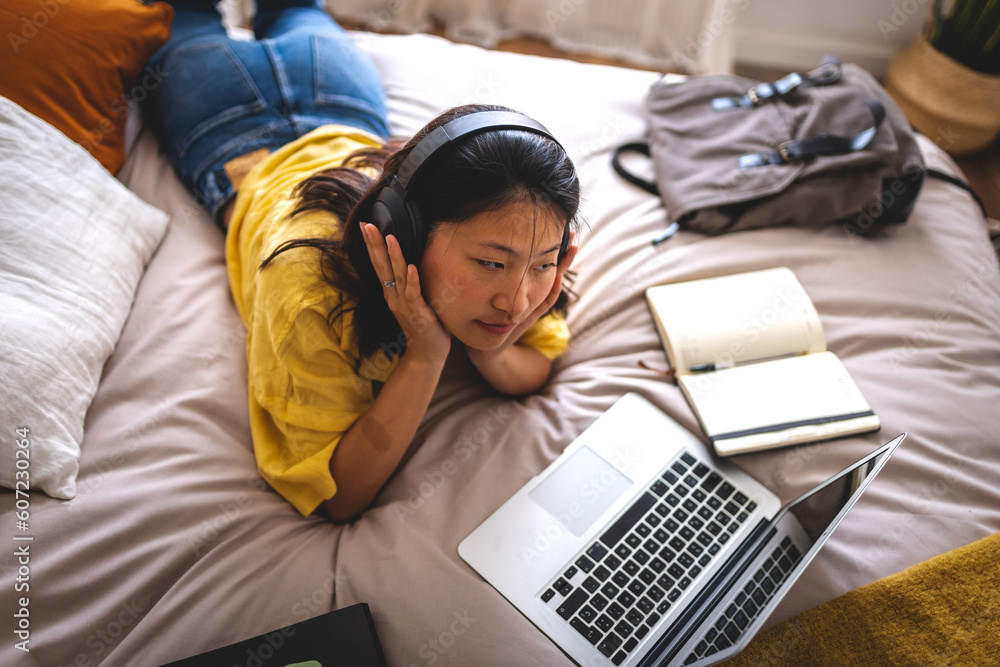Asian college student at home lying on bed using laptop attending online university classes listening with headphones. - obrazy, fototapety, plakaty 