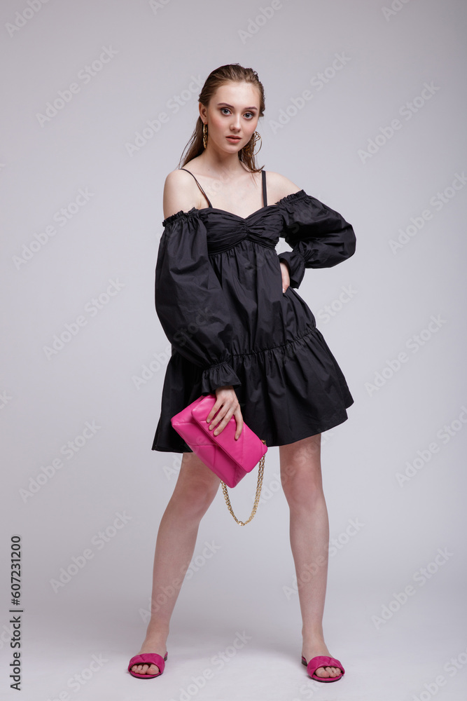 Fototapeta premium High fashion photo of beautiful elegant young woman in a pretty black dress, pink handbag, clutch posing over white, soft gray background. Studio Shot, portrait.