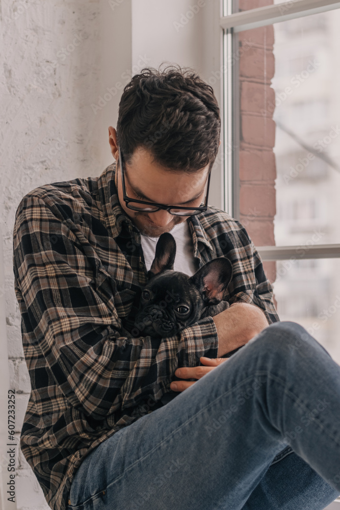 Obraz premium A young positive man is embracing a puppy, a black French Bulldog, sitting on the windowsill in an apartment.The concept of care, training,raising of animals.