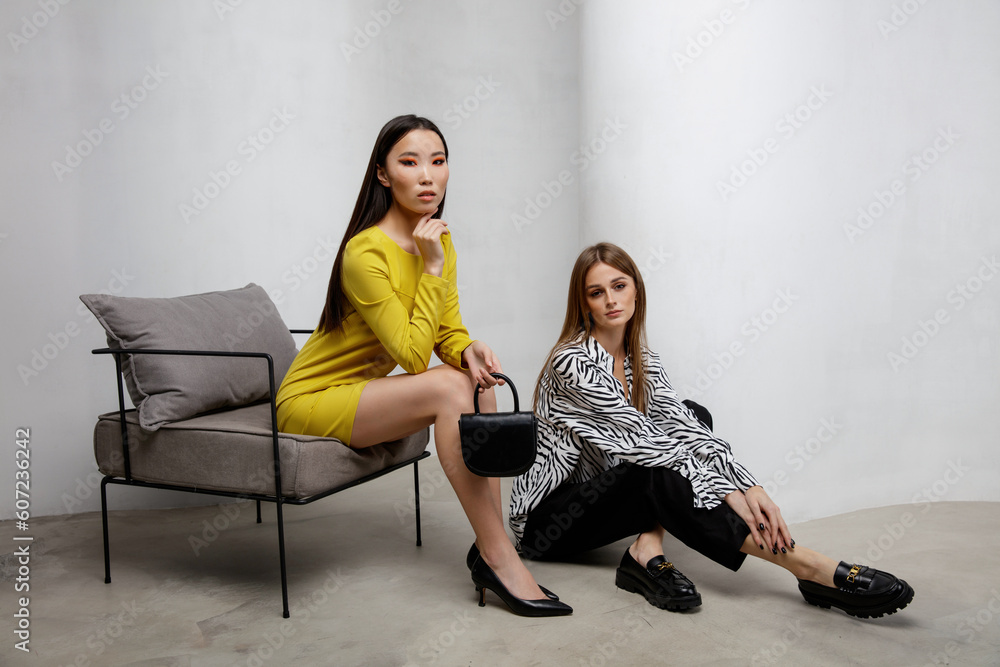 Two fashion models in short yellow dress, handbag, clutch, zebra shirt, pants, trousers. Beautiful young women. Textured white wall. Asian, european girls Two fashion models in short yellow dress, handbag, clutch, zebra shirt, pants, trousers. Beautiful young women. Textured white wall. Asian, european girls