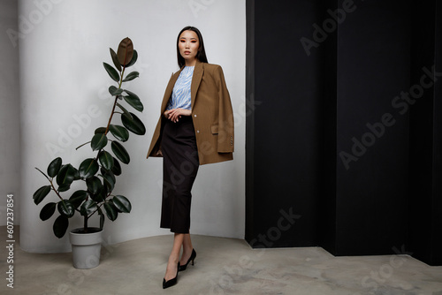 High fashion photo of beautiful elegant young asian woman in pretty brown beige jacket, blazer, blue top, long skirt posing on textured black and white wall, tall flower. Slim figure