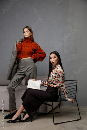 Two fashion models in orange beige outfits. Sweater, shirt, black skirt, gray pants, trousers, clutch, handbag. Beautiful young women. Asian and european girls posing on textured wall, tall flower