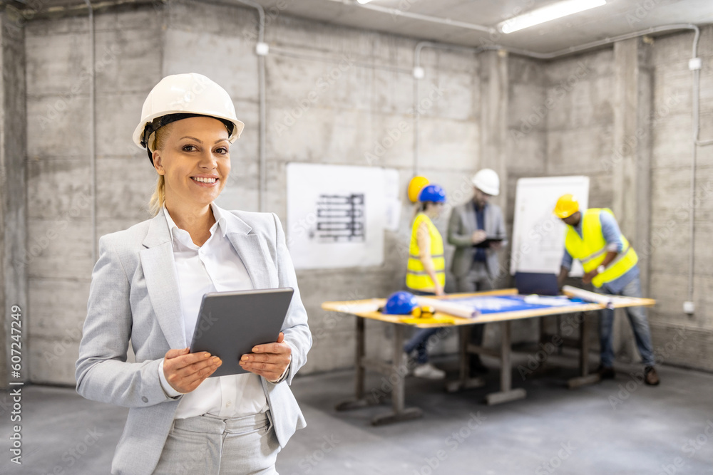 Female structural engineer with digital tablet standing at construction site while her colleagues brainstorming in background. Female structural engineer with digital tablet standing at construction site while her colleagues brainstorming in background.