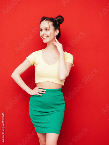 Young beautiful smiling female in trendy summer yellow t-shirt and green skirt. Carefree woman with two horns hairstyle posing near red wall in studio. Positive model having fun. Cheerful and happy