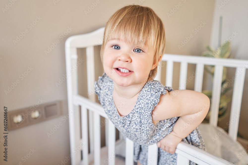 Happy beautiful baby girl one and half standing in the crib. Smiling 18 months child playing in sunny bedroom. Baby cot in the parent's bedroom. Happy beautiful baby girl one and half standing in the crib. Smiling 18 months child playing in sunny bedroom. Baby cot in the parent's bedroom.