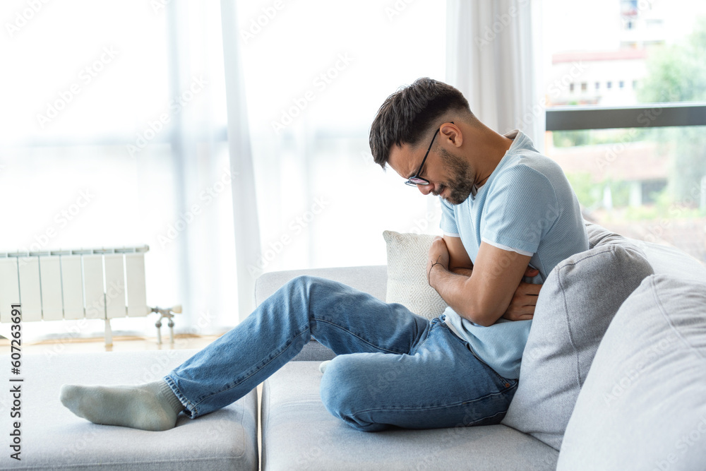 A man suffers from abdominal pain while sitting at home on the couch. Young man suffering from abdominal pain sitting on the couch at home. A man suffers from abdominal pain while sitting at home on the couch. Young man suffering from abdominal pain sitting on the couch at home.
