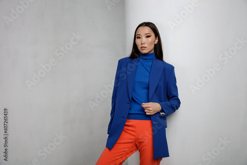 High fashion photo of a beautiful elegant young asian woman in pretty blue jacket, blazer, orange pants, trousers. White textured rounded wall