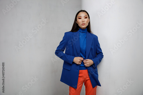 High fashion photo of a beautiful elegant young asian woman in pretty blue jacket, blazer, orange pants, trousers. White textured rounded wall