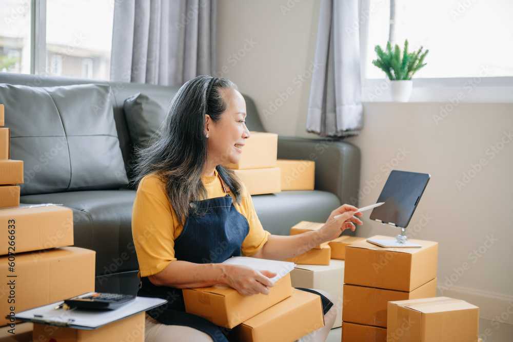 Mature business owner woman prepare parcel box and standing check online orders for deliver to customer on tablet, laptop Shopping Online Mature business owner woman prepare parcel box and standing check online orders for deliver to customer on tablet, laptop Shopping Online