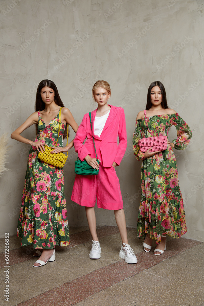 Three fashion models. Asians in identical looks, green dress with floral pattern, handbag, clutch. Blonde in pink fuchia suit, jacket, blazer, shorts. Beautiful young women. Sandy beige textured wall