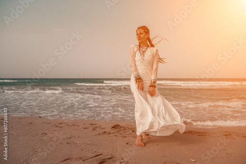 Model in boho style in a white long dress and silver jewelry on the beach. Her hair is braided, and there are many bracelets on her arms. © svetograph