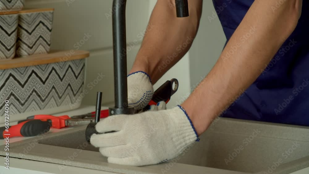 Close-up of male hands in protective gloves repairing kitchen faucet with adjustable wrench while providing plumbing service indoors.