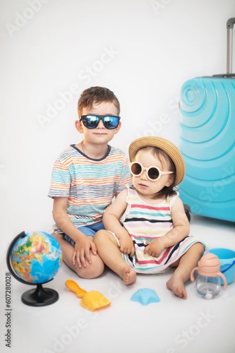 A cute boy and a girl are sitting on a light background in striped bright clothes near a blue suitcase with sand toys and a globe in anticipation of a trip, vacation with children