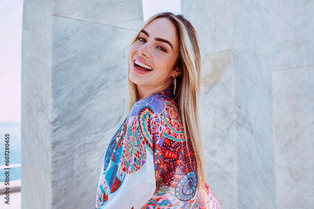 Young charming blond latin american woman smiles broadly, enjoys summer vacations. Female outdoors wearing colorful smock with marble column background.