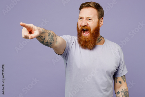 Young mad furious indignant redhead bearded man wear violet t-shirt casual clothes point index finger aside scream isolated on plain pastel light purple background studio portrait. Lifestyle concept. photo