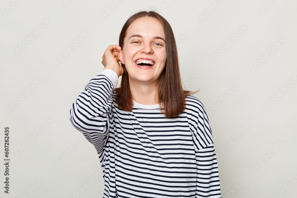 Beautiful optimistic woman with brown hair wearing striped shirt standing isolated over gray background being in good mood flirting keeps hand on hair. Beautiful optimistic woman with brown hair wearing striped shirt standing isolated over gray background being in good mood flirting keeps hand on hair.