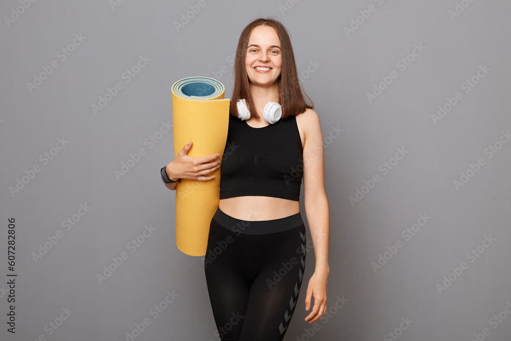 Training yoga lifestyle. Young beautiful Caucasian woman wearing sportswear holding yoga mat isolated on gray background, preparing for exercise workout in gym. Training yoga lifestyle. Young beautiful Caucasian woman wearing sportswear holding yoga mat isolated on gray background, preparing for exercise workout in gym.