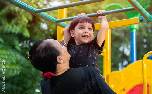 Portrait of a funny little Asian boy hangs by horizontal bar with mother on playground in the sunny day.