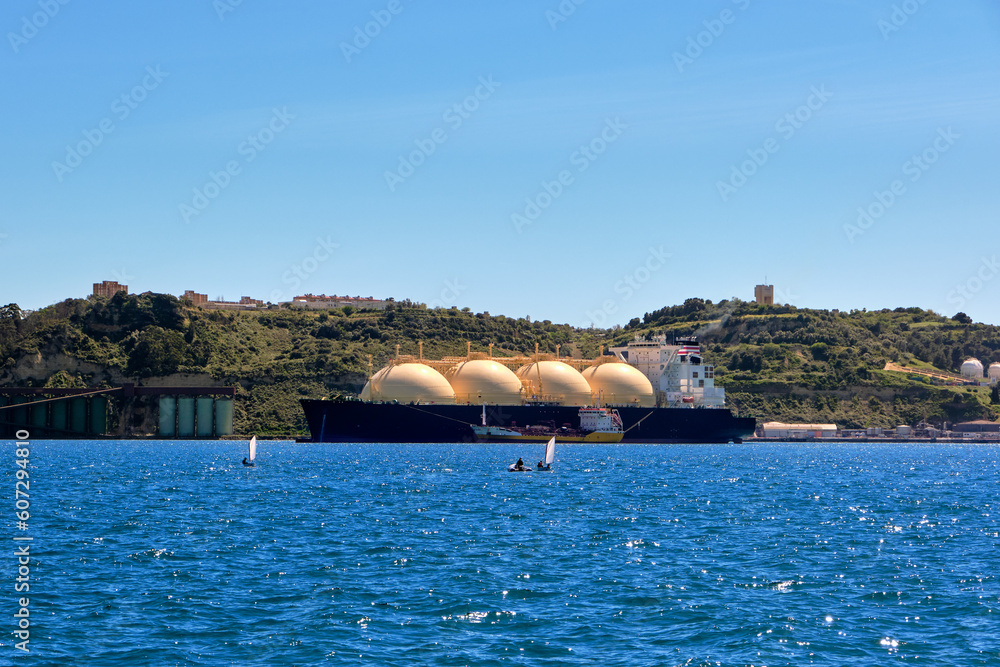 LNG tanker moored by industrial pier and green hill on sunny day