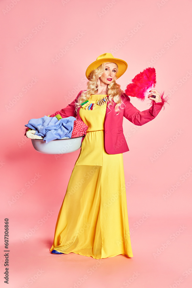 Full-length portrait of stylish, beautiful senior lady, woman in bright yellow dress holding laundry against pink studio background. Concept of beauty, fashion, human emotions, lifestyle. Ad