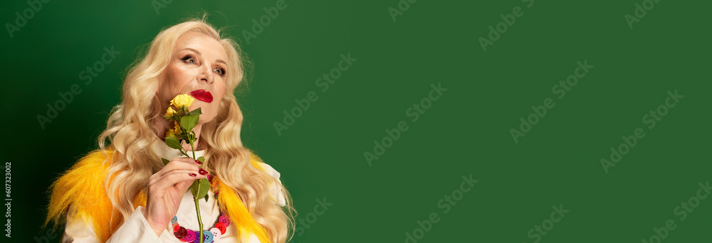 Portrait of beautiful lady with long blonde hair and makeup holding yellow roses against green studio background. Concept of beauty, fashion, human emotions, lifestyle. Banner. Copy space for ad