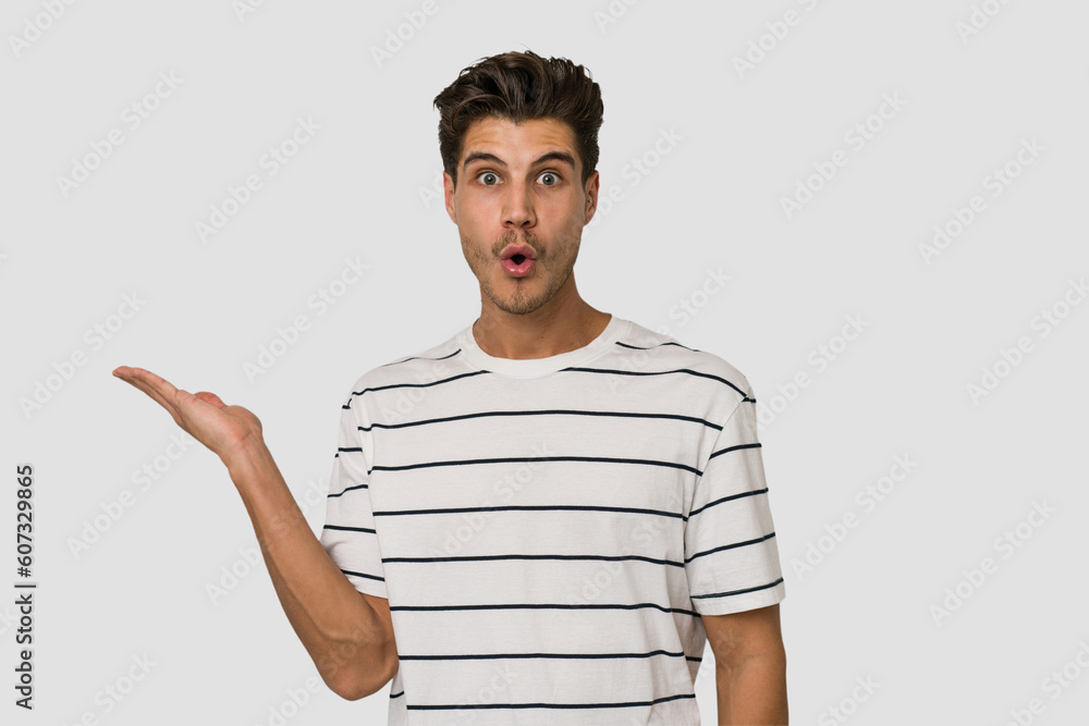 Young handsome caucasian man isolated on white background impressed holding copy space on palm. Young handsome caucasian man isolated on white background impressed holding copy space on palm.